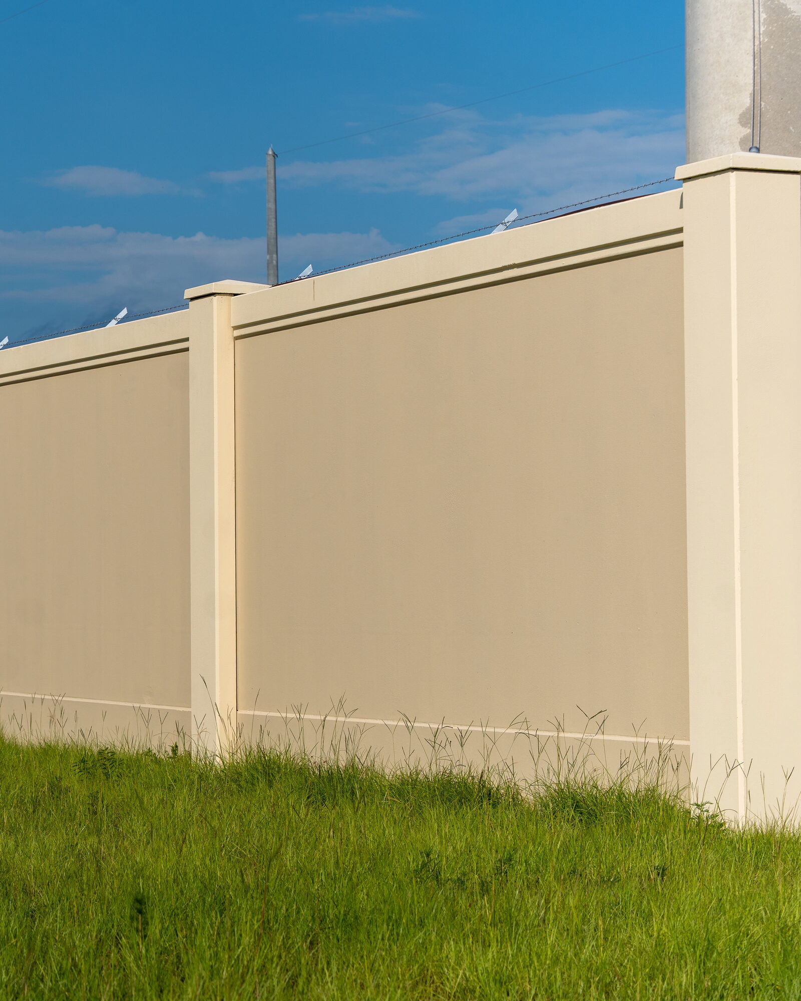 Eye-level run of tan precast concrete wall panels set between capped posts, grass in the foreground and a concrete pole rising behind the corner post.