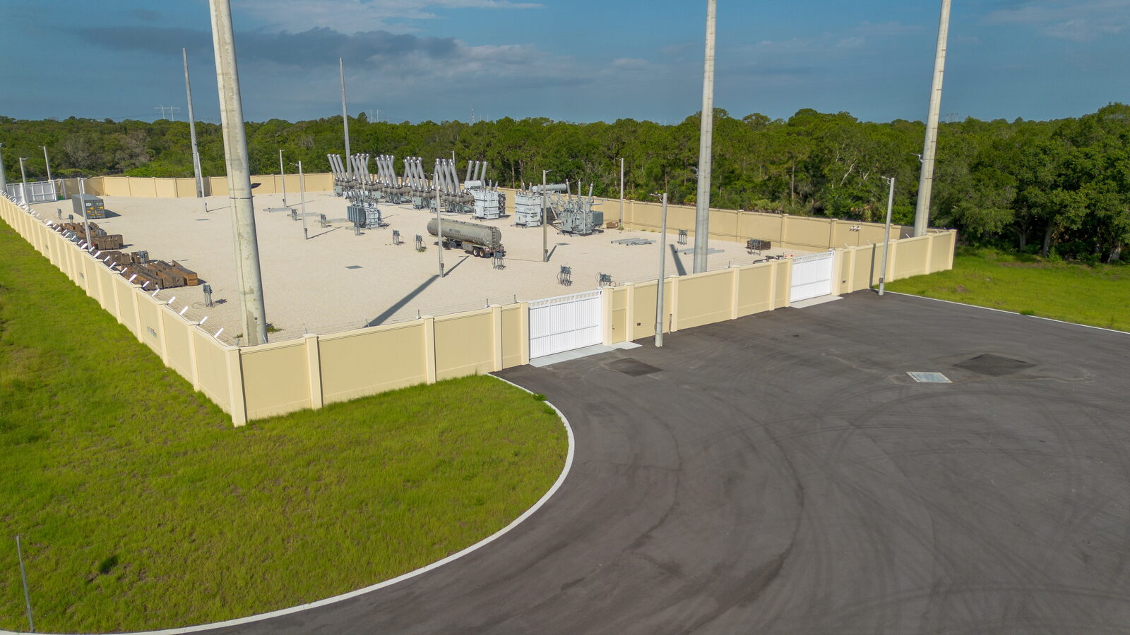 Drone view of a substation enclosed on all four sides by a tan precast concrete panel wall, two white gates opening onto a fresh asphalt apron, transformers and bus structures inside.