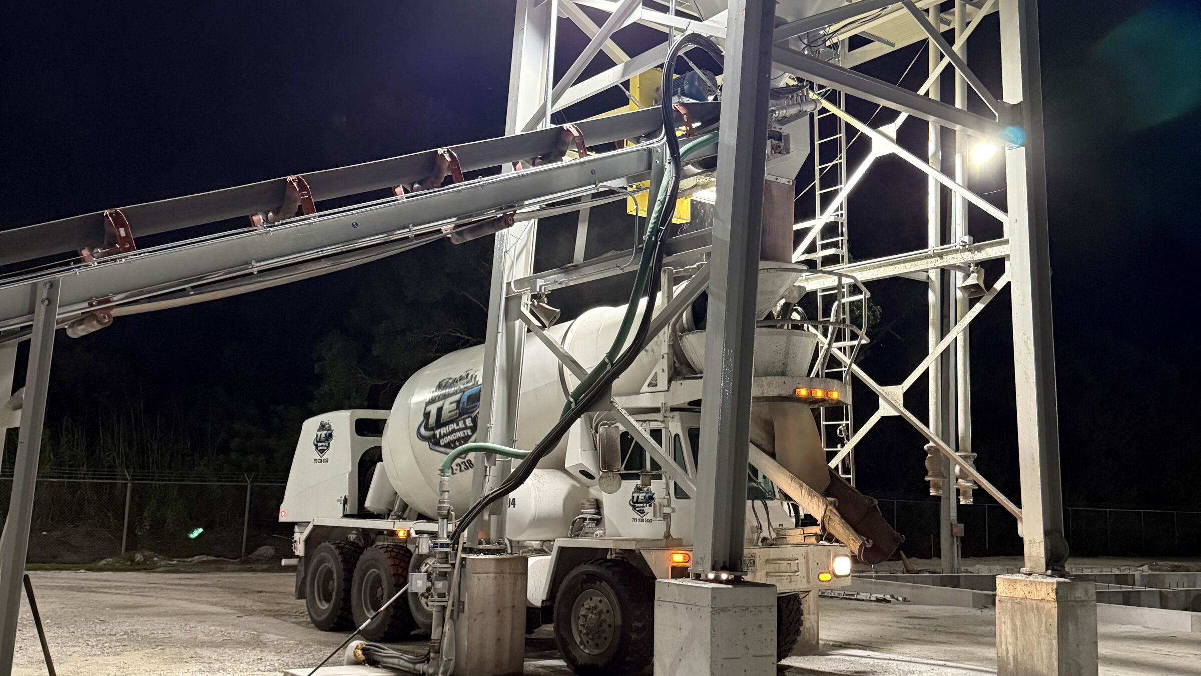 A white Triple E Concrete mixer truck charging under the floodlit batch plant at night in Indiantown, Florida, the aggregate conveyor running overhead against a black sky.
