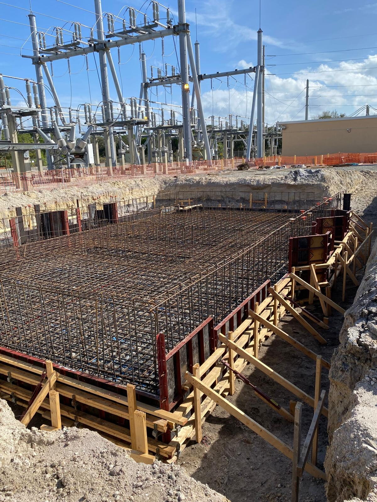 A dense rebar cage for a substation foundation, tied inside the excavation with substation steel and blue sky behind.